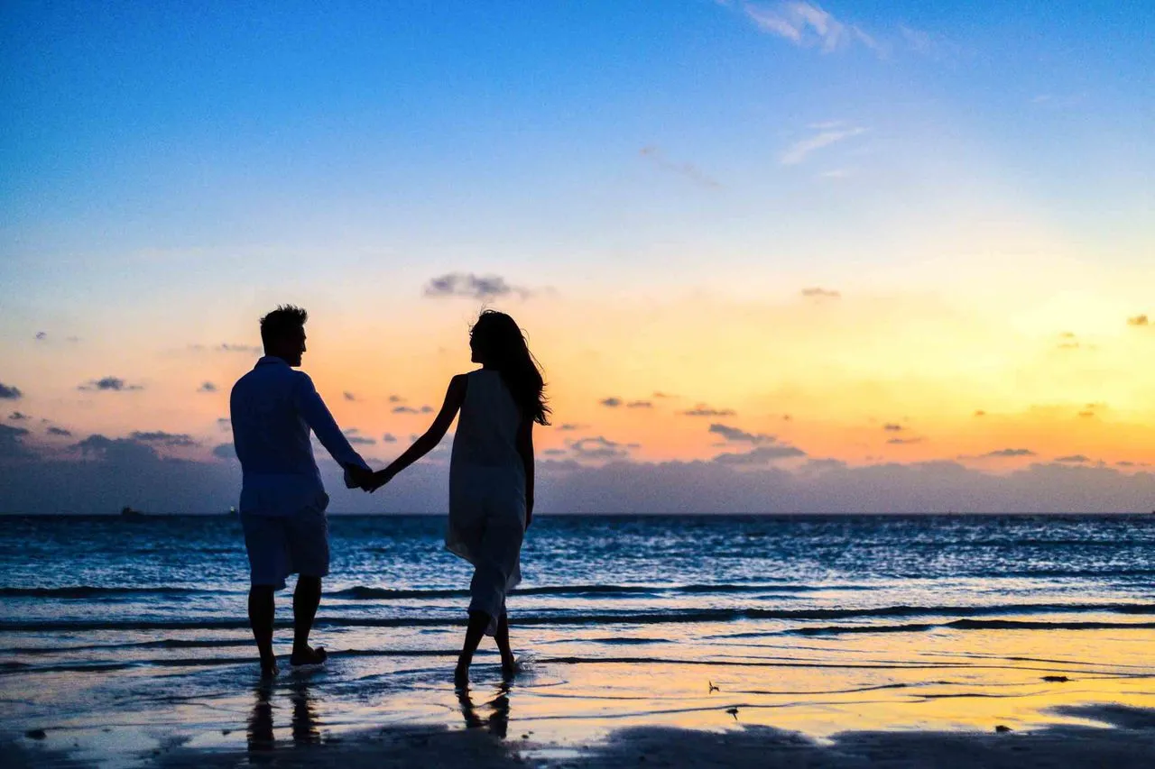A couple walking along a white-sand beach at golden hour with turquoise water stretching to the horizon